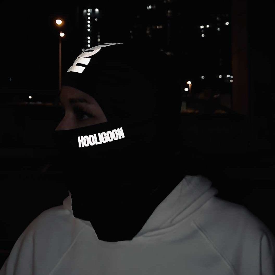 Person wearing a reflective 'Hooligoon' mask at night with city lights in the background