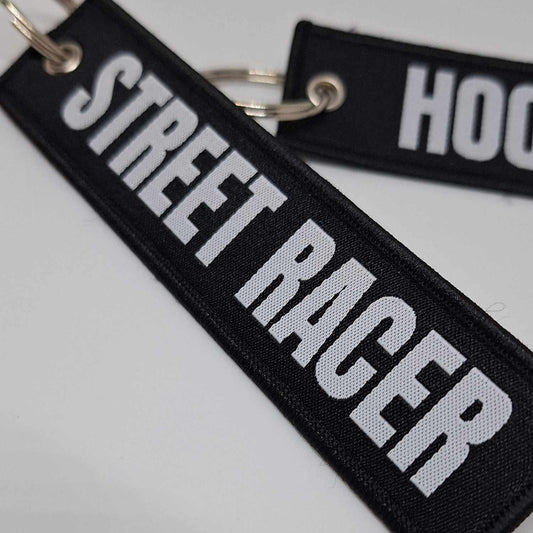 Two black key tags with white text, one reading 'STREET RACER' and the other 'HOOLIGOON'. They are laid out on a white surface.