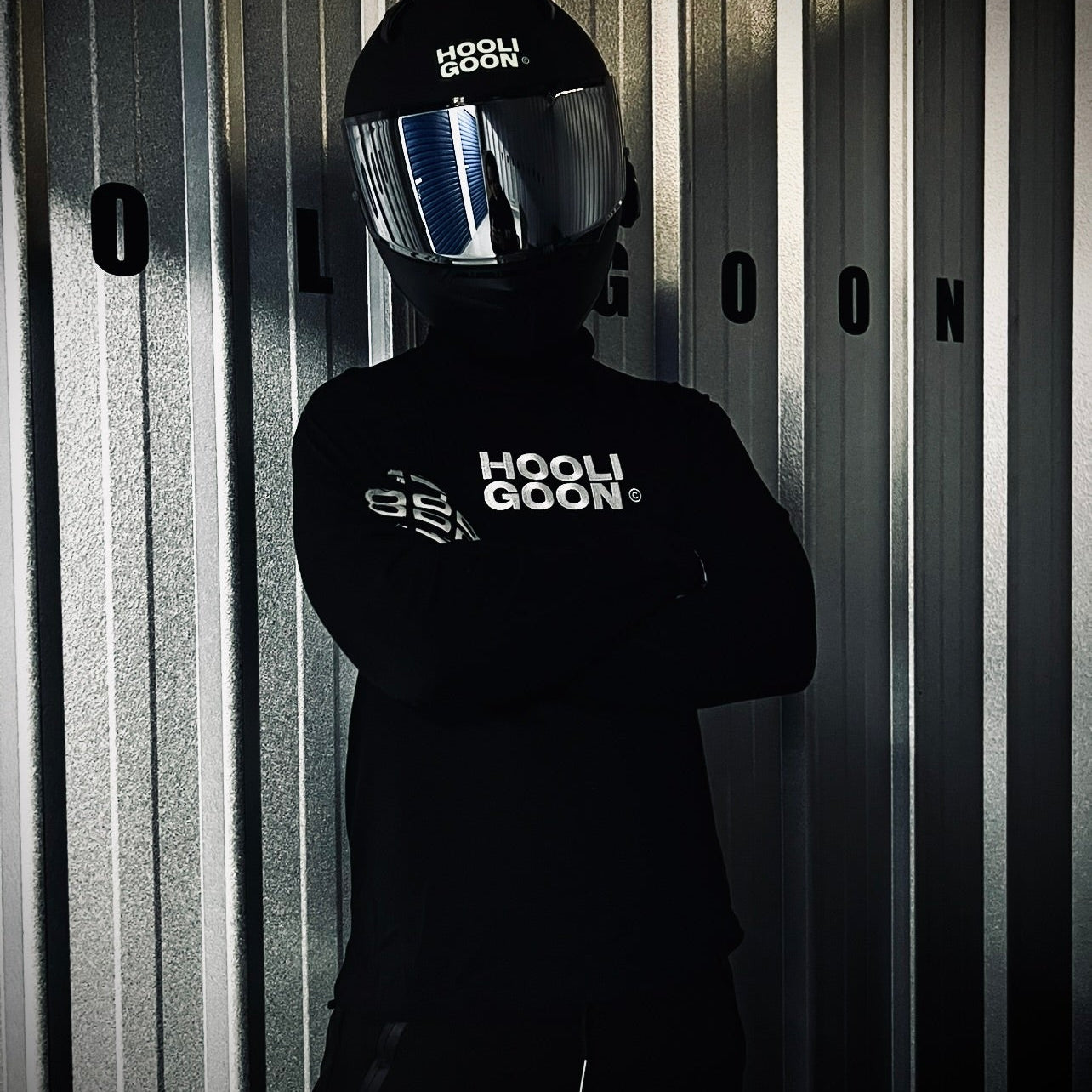 Person wearing a black 'HOOLIGOON' helmet and jacket against a corrugated metal background.