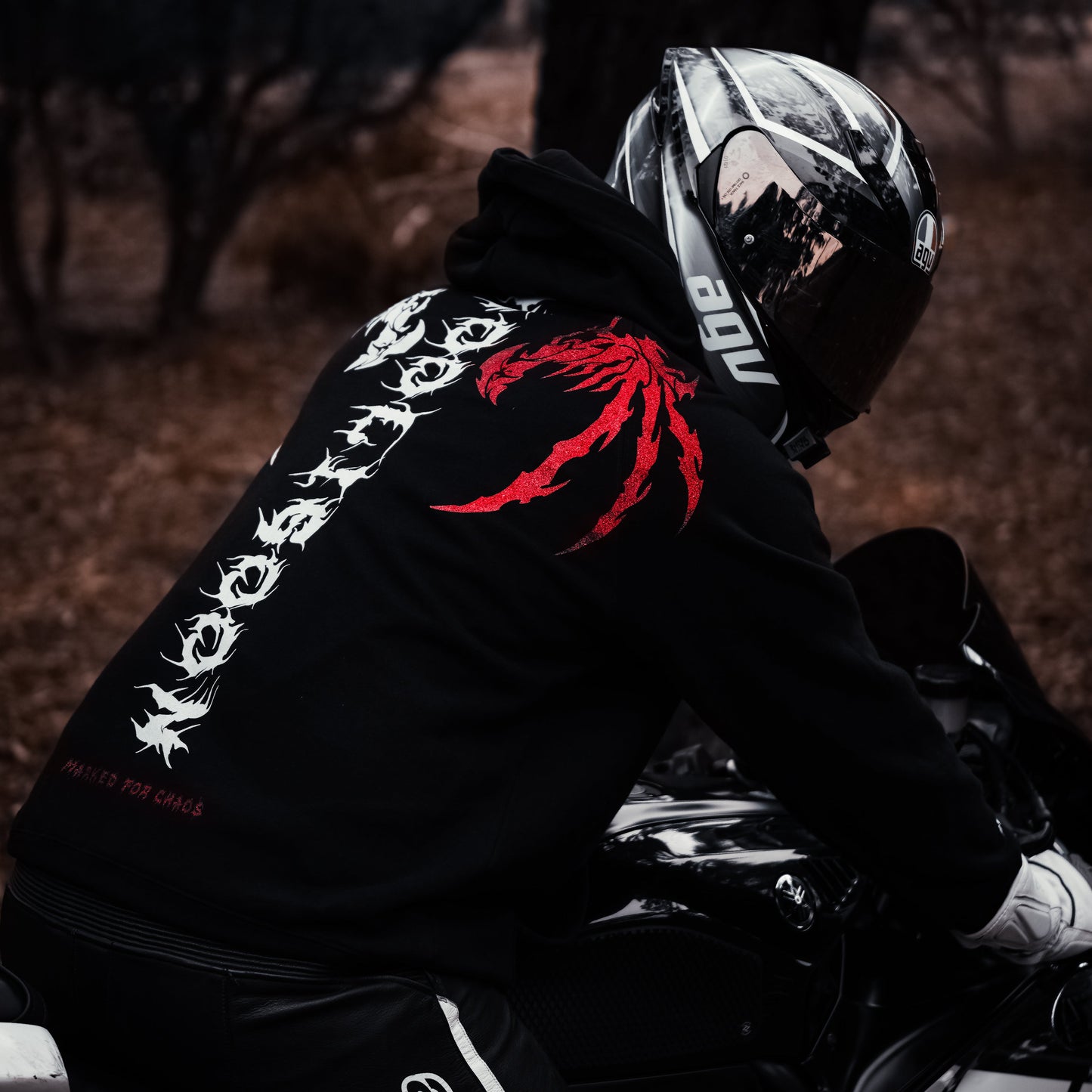 Person wearing a black hoodie with red and white designs, sitting on a motorcycle in a dark setting.