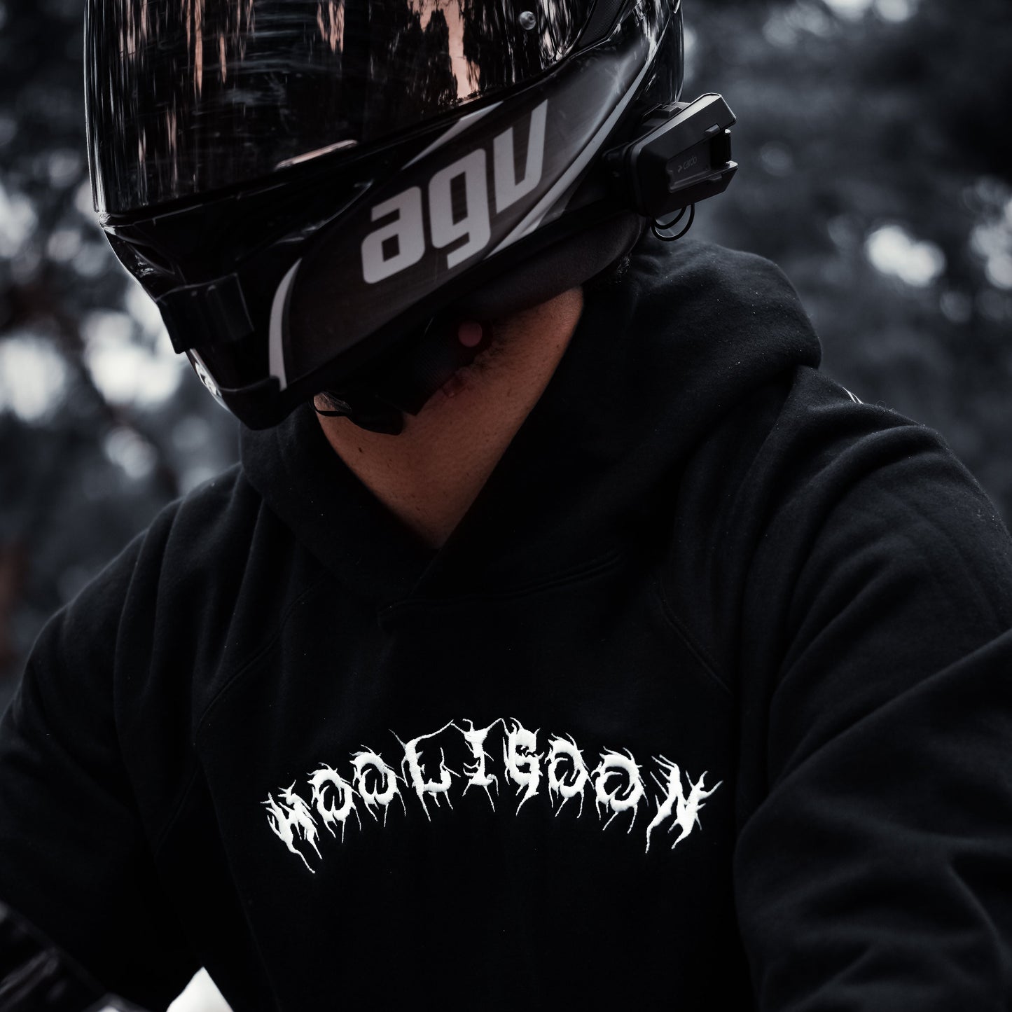 Person wearing a black hoodie with 'Hooligoon' text and an AGV helmet, sitting on a motorcycle.