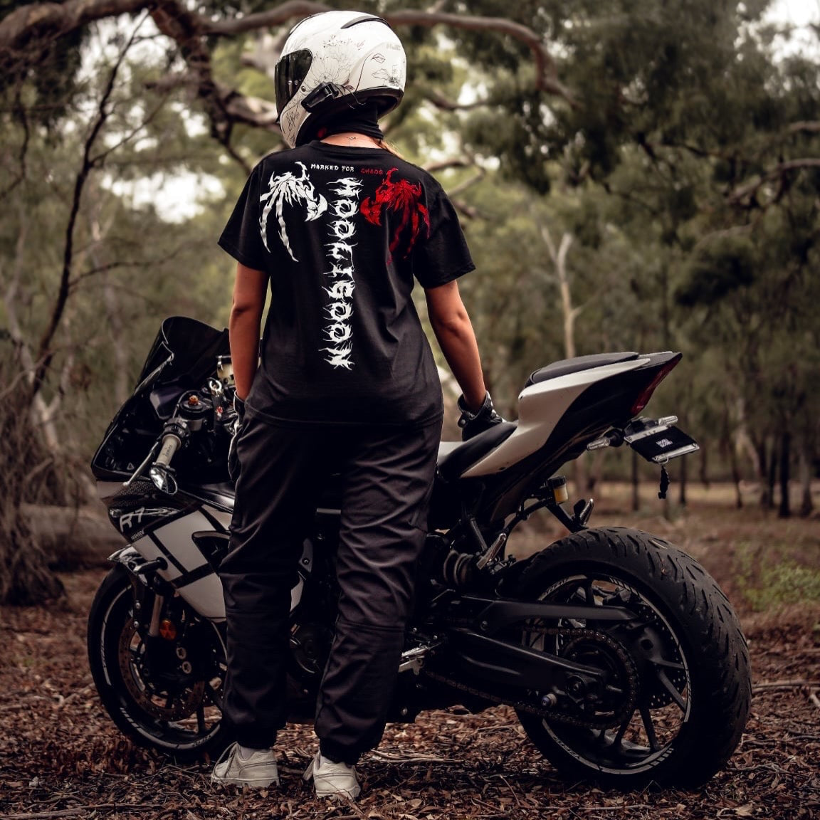 Person wearing a black t-shirt with visible branding and text, standing next to a motorcycle in a forest setting.