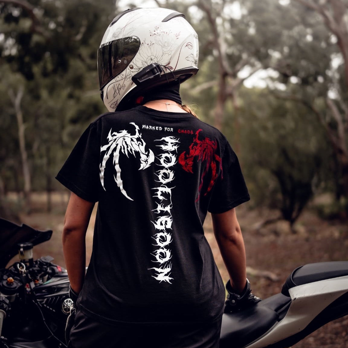 Person wearing a black t-shirt with white text and graphics, standing next to a motorcycle in a forest setting.