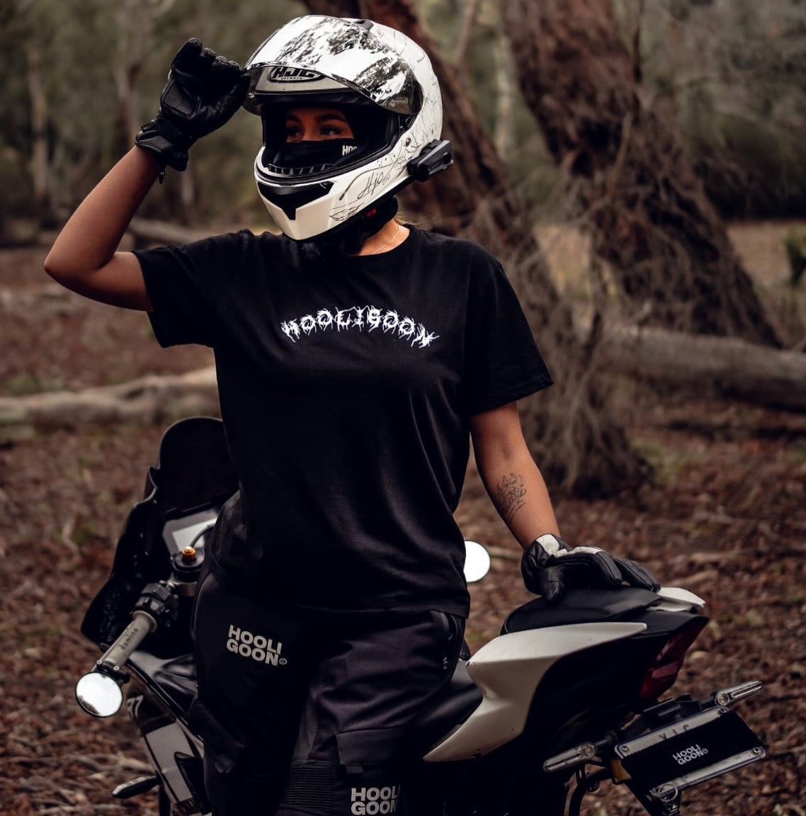 Person wearing a black 'Hooligoon’ t-shirt and helmet, standing next to a motorcycle in a forest setting.