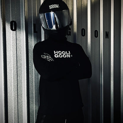 Person wearing a black 'HOOLIGOON' helmet and jacket against a corrugated metal background.