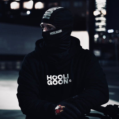 Person wearing a dark hoodie with 'HOOLIGOON' text, sitting against a blurred cityscape background.
