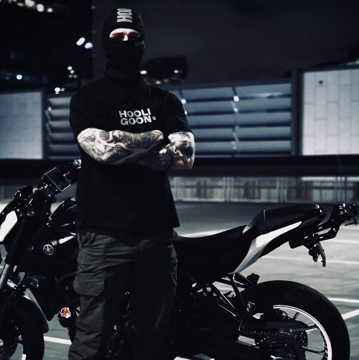 Person wearing a black 'Hooligoon' shirt standing next to a motorcycle in an urban setting.