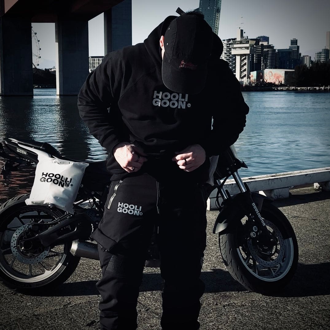 Person wearing a black hoodie with 'HOOLIGOON' branding, standing next to a motorcycle by a waterfront with city skyline in the background.