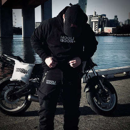 Person wearing a black hoodie with 'HOOLIGOON' branding, standing next to a motorcycle by a waterfront with city skyline in the background.