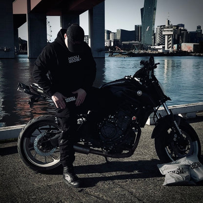 Person standing next to a motorcycle by a waterfront with city skyline in the background